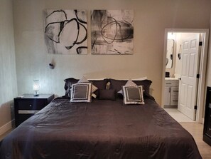 Quarto com cama grande, duas pinturas abstratas na parede, mesa de cabeceira com abajur e porta de acesso a um banheiro.
