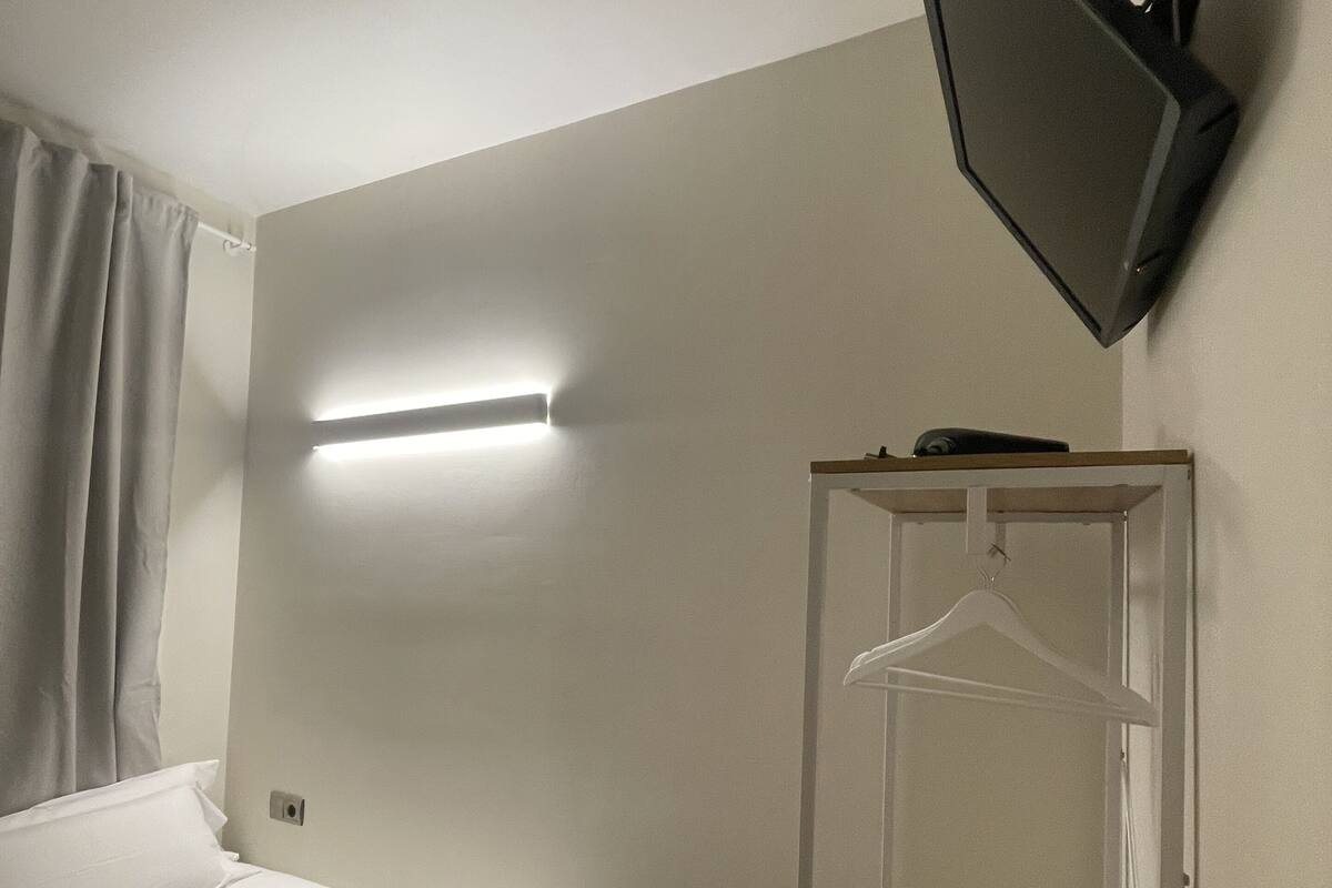 A hotel room with a bed, a hanging clothes rack, a small wooden stool, and a mounted television.