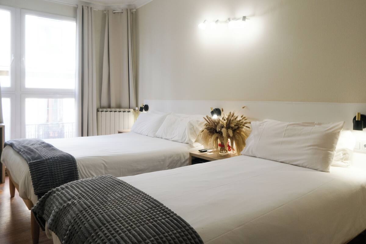 A hotel room with two beds, a bedside table with a lamp, and a window with curtains.