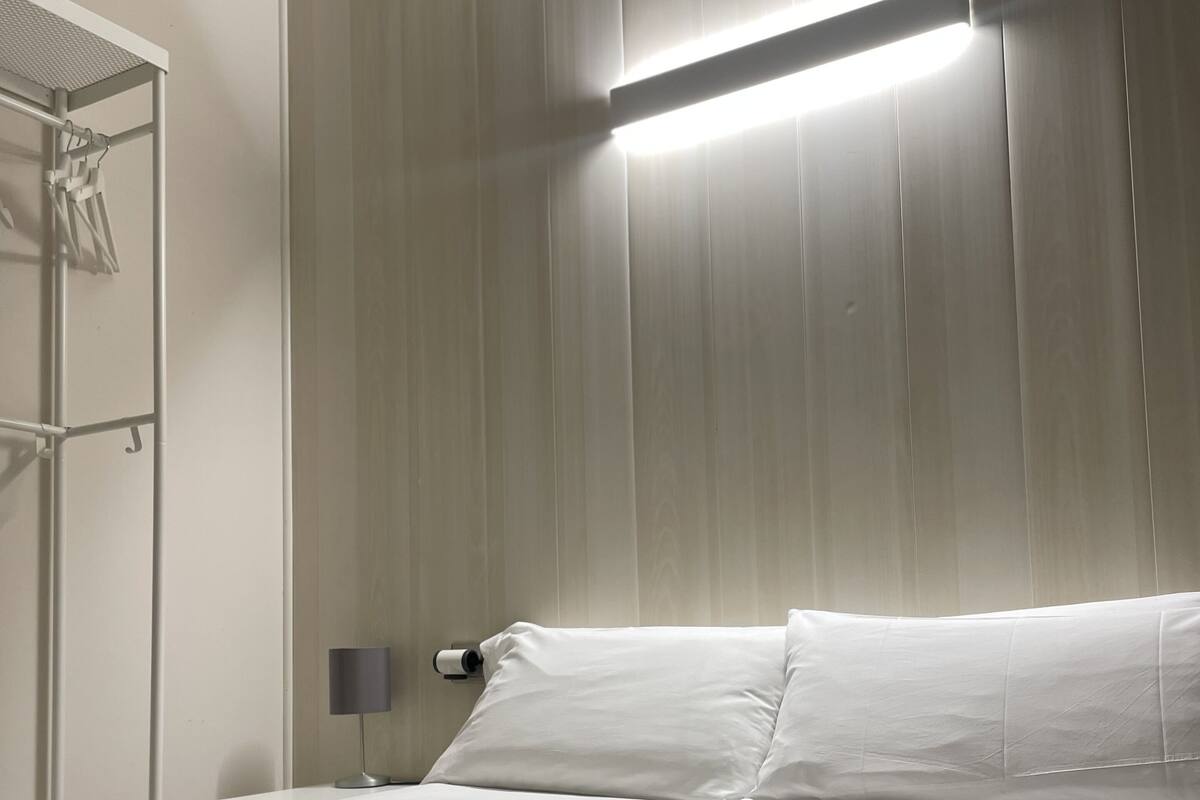 A neatly made bed with white linens and a folded towel, a bedside lamp, and a wall-mounted shelf with a towel rack.
