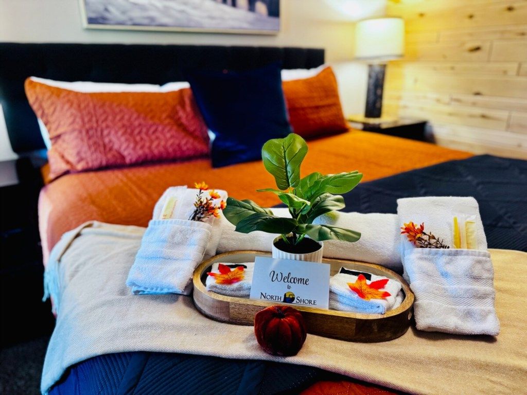Ett snyggt uppdukat välkomstbricka med handdukar och en växt på sängen, en skylt med texten ”Välkommen till North Shore” och en röd dekorativ detalj.