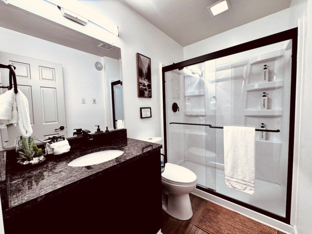 A modern bathroom with a large mirror, a double sink, a toilet, and a glass-enclosed shower area.