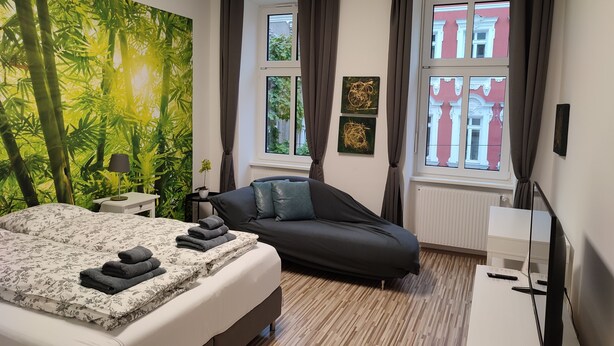 A bedroom with a bed, a sofa, a window with curtains, a wall-mounted TV, and a large green wall mural.