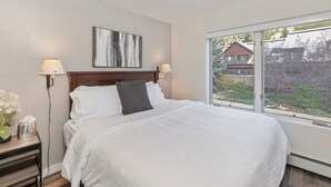 Ein ordentlich gemachtes Bett mit weißen Leinen und einem grauen Kopfkissen, ein Nachttisch mit Lampe und ein Fenster mit Blick auf ein Haus und Bäume.