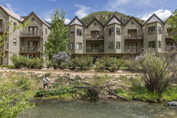 Un hôtel à plusieurs étages doté de balcons, situé en bord de rivière, avec une chaîne de montagnes en arrière-plan.