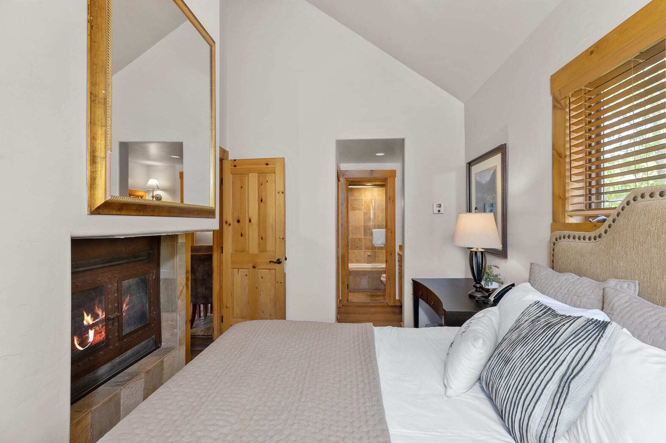 A bedroom with a fireplace, wooden doors, a bed with pillows, a nightstand with a lamp, and a window with blinds.