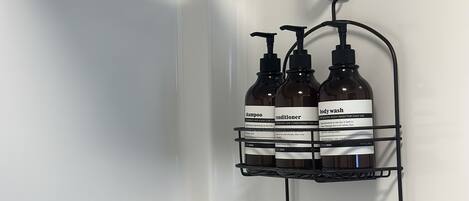 Une salle de bain avec une douche, une baignoire blanche, un porte-serviettes et deux étagères où sont rangées des bouteilles de gel douche et d’après-shampoing.