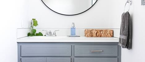 A bathroom with a grey vanity, a round mirror, a plant, and a towel rack.