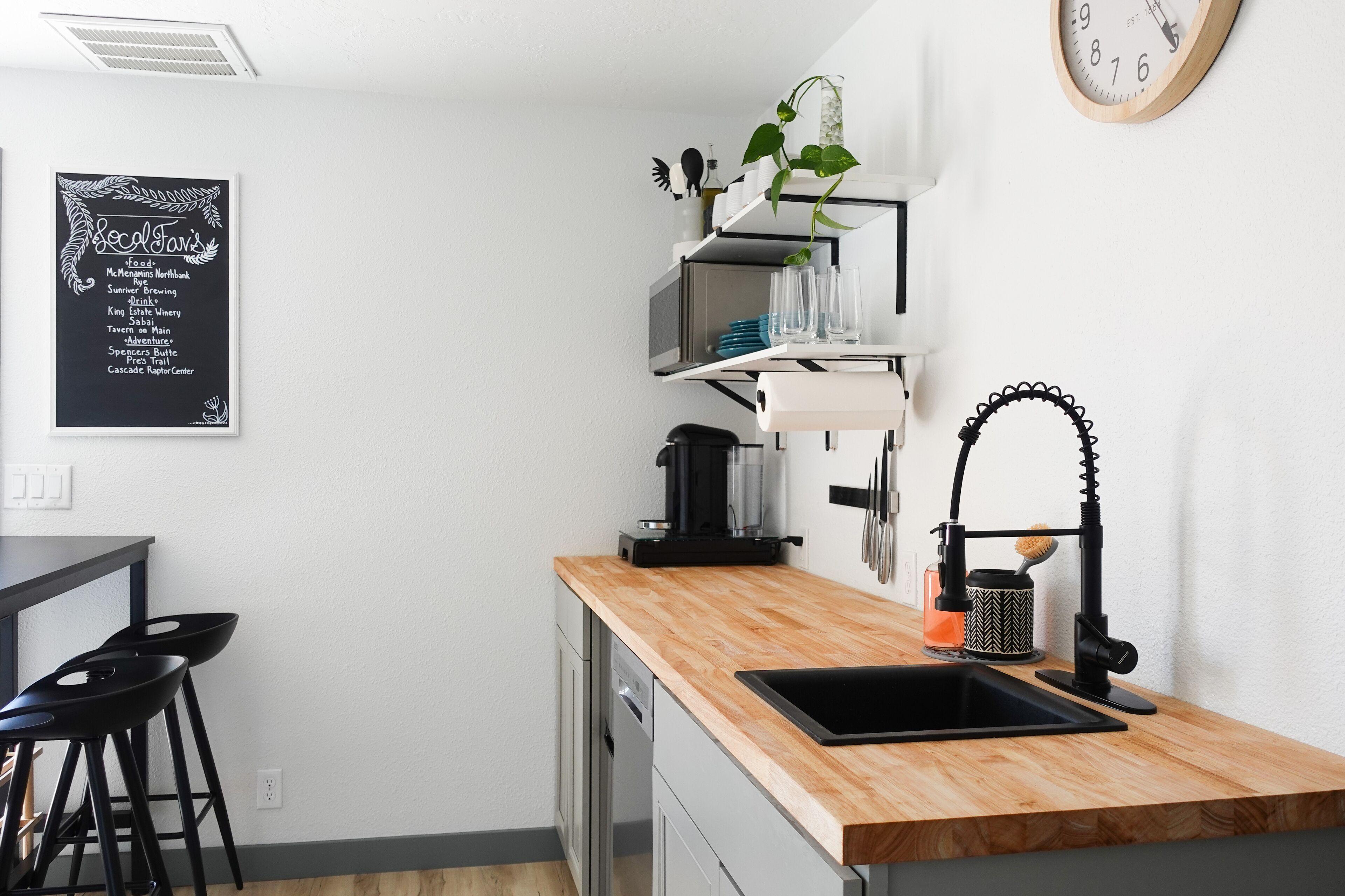 A modern kitchen with a wooden countertop, a black sink, and a coffee machine. There is a chalkboard menu on the wall listing food and drink options.