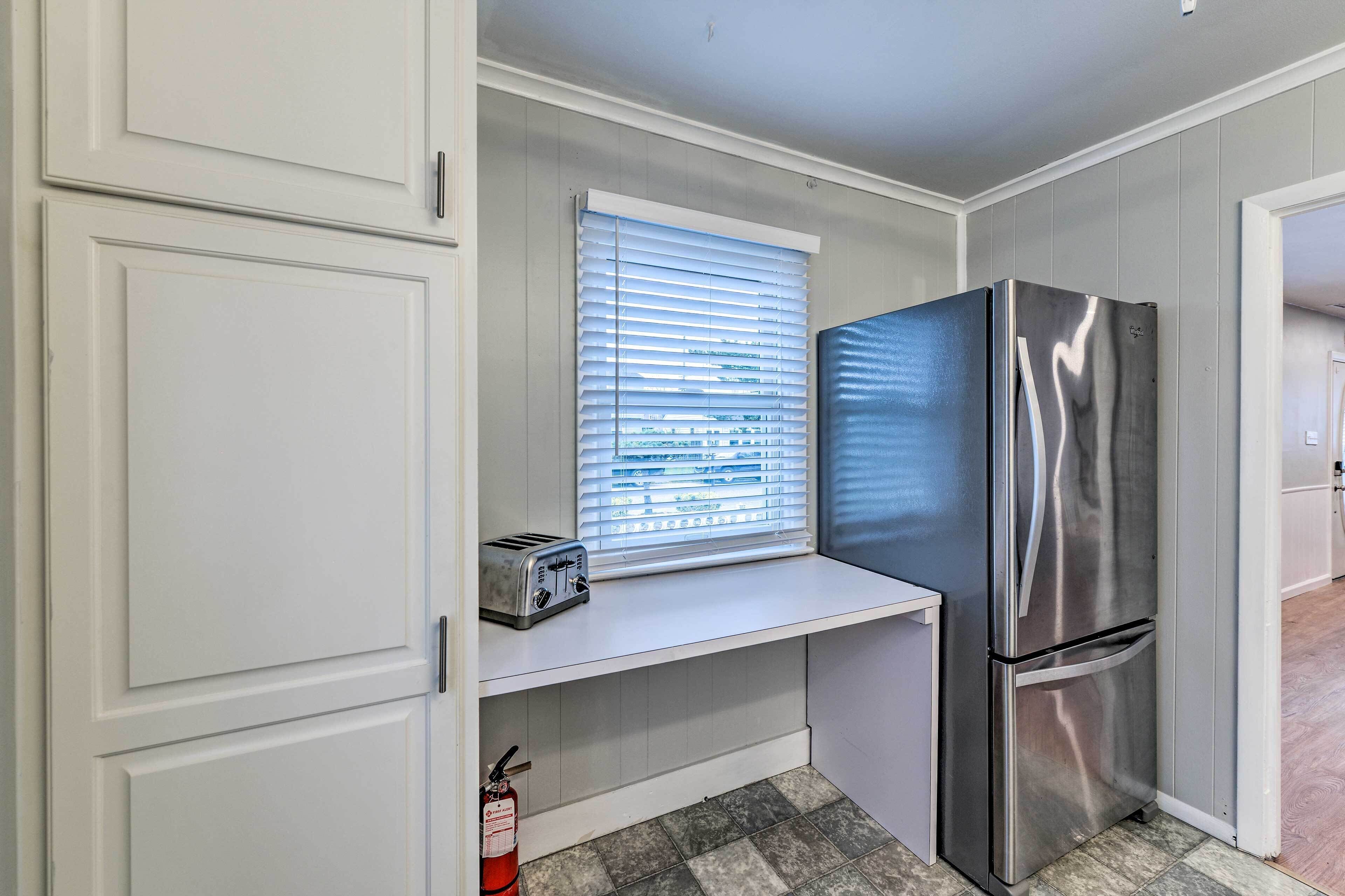A compact kitchen with white cabinets, a stainless steel refrigerator, a window with blinds, and a toaster on a shelf.