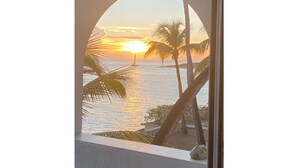 A sunset view through a circular window overlooking a beach with palm trees and a sailboat. - Oceanfront, 4 balcony condo: Stunning Sunsets (Sint Maarten)