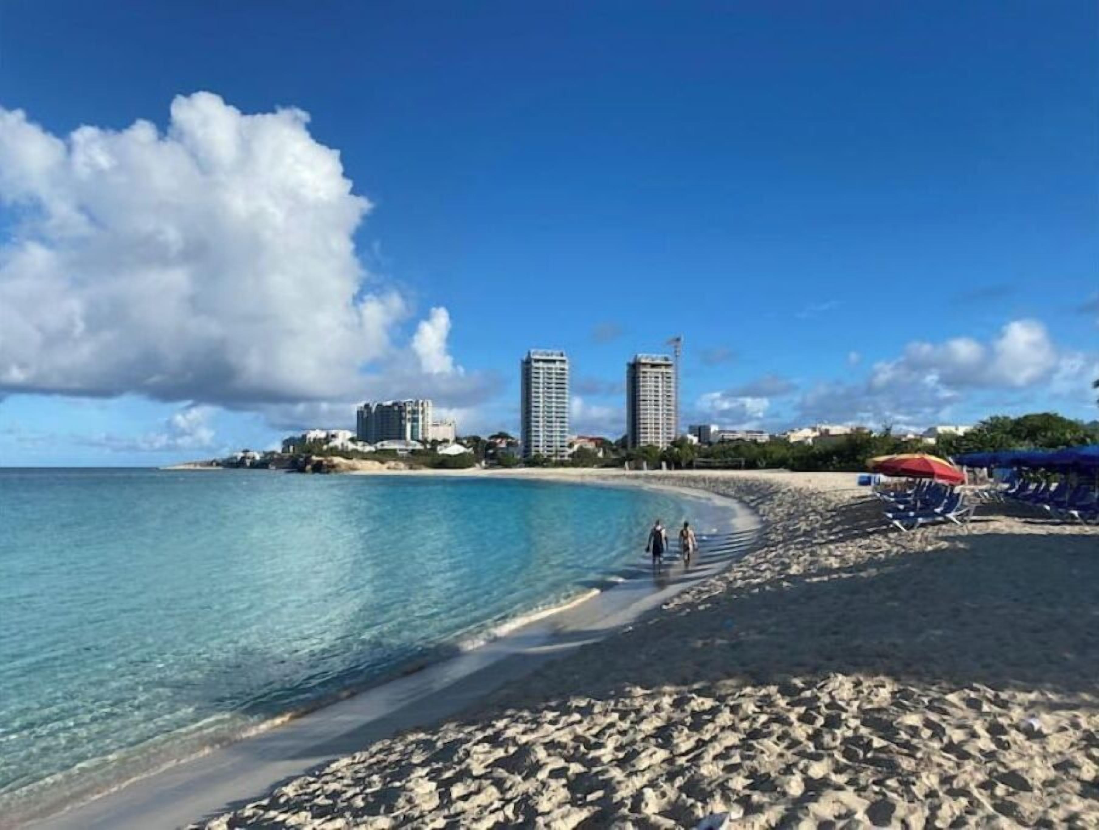 Ein Strand mit klarem türkisblauem Wasser, einem Sandufer und einigen Spaziergängern. Im Hintergrund sind hohe Gebäude zu sehen, und ein roter Sonnenschirm steht im Sand.
