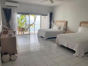 A hotel room with two beds, a large window with a view of palm trees and the sea, a ceiling fan, and a small table with a basket and two decorative vases. - Oceanfront, 4 balcony condo: Stunning Sunsets (Sint Maarten)