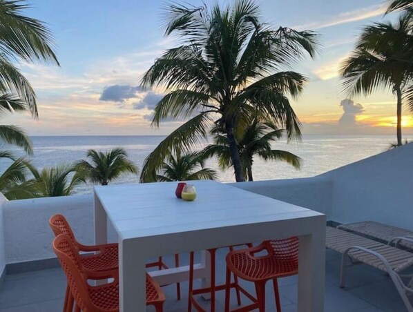 A balcony with a white table and orange chairs, overlooking the ocean at sunset. - Oceanfront, 4 balcony condo: Stunning Sunsets (Sint Maarten)