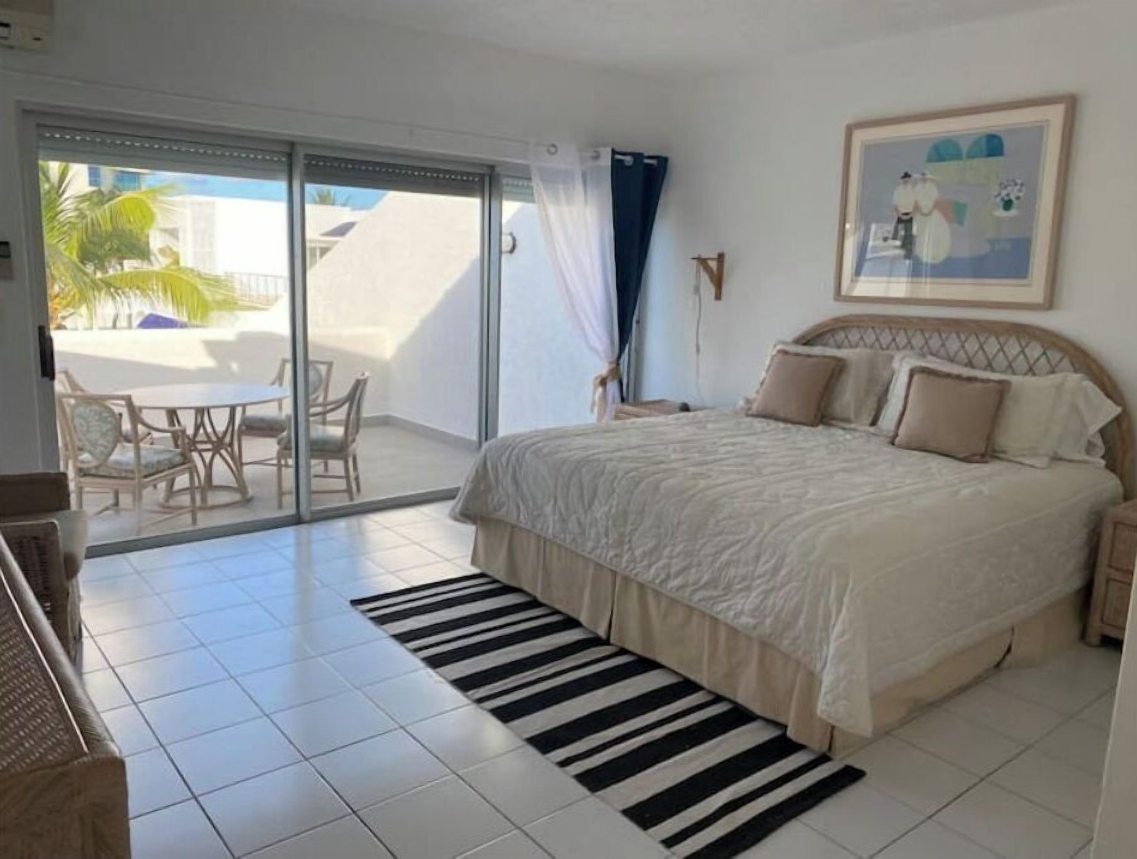 A bedroom with a large bed, a view of the outside, a dining area with chairs, and a sliding glass door to the balcony.
