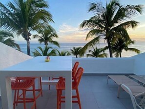 A terrace with a white table and red chairs, overlooking the ocean with palm trees. - Oceanfront, 4 balcony condo: Stunning Sunsets (Sint Maarten)