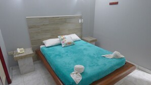 Un lit bien fait, recouvert d’une couvre-lit turquoise, avec deux oreillers et deux serviettes pliées au pied du lit. On y trouve une table de chevet en bois avec un téléphone et une petite table placée à côté du lit.