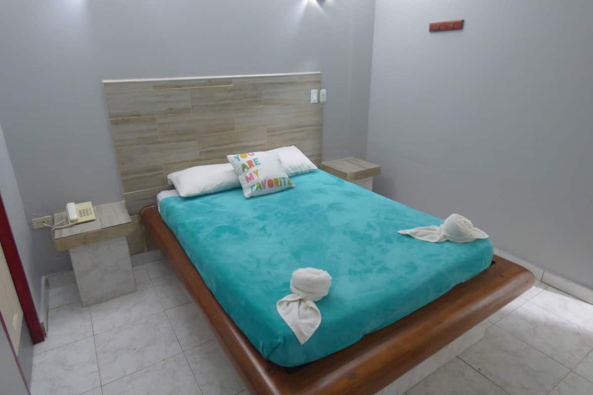 A neatly made bed with a teal bedspread, two pillows, and two towels folded at the foot of the bed. There is a wooden nightstand with a phone and a small table beside the bed.