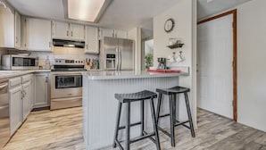 Une cuisine moderne avec des armoires blanches, des appareils en acier inoxydable, un grand îlot central doté de tabourets et une horloge au mur.