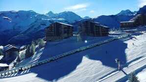A snow-covered mountain landscape with a bright sun, a ski slope, and several buildings with balconies. - Charming snow front studio (Auris)