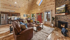 A spacious living room with a fireplace, a cozy fireplace area, and a kitchen with wooden cabinets and a stainless steel refrigerator. - Family Friendly Cabin, Mountain Views, Easy Roads, Pool, Hot Tub, Game Room (Gatlinburg)