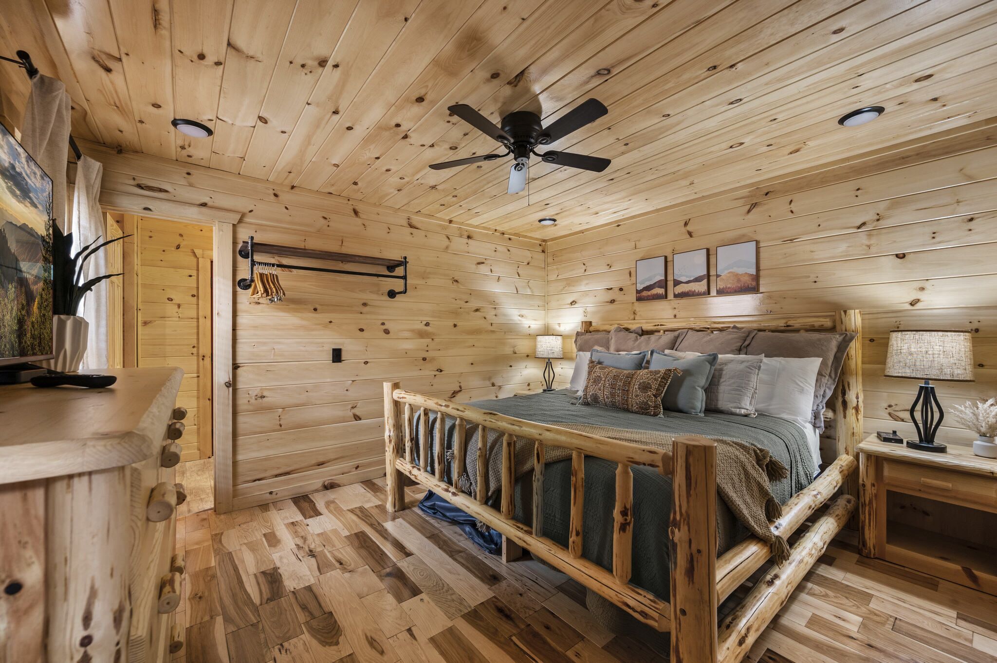 Ein Schlafzimmer einer Holzhütte mit einem Bett, einem Deckenventilator, einem Nachttisch und einem an der Wand befestigten Kleiderhaken.