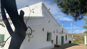 A white building with a green door and window shutters, a small garden area, and a view of hills in the background.
