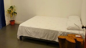 Una cama pequeña y ordenada con colcha y almohadas blancas, una planta en maceta y un taburete de madera en una habitación sencilla con paredes lisas y suelo de baldosas.