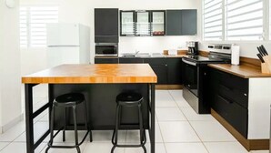 A modern kitchen with black cabinets, a wooden table, two black stools, a refrigerator, an oven, a sink, and a window with blinds. - Home in the heart of the city (Guaynabo)
