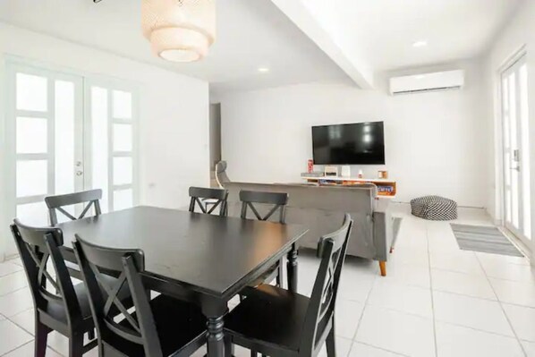Un comedor moderno con una mesa y sillas negras, un televisor de pantalla plana montado en la pared y una zona de cocina al fondo.