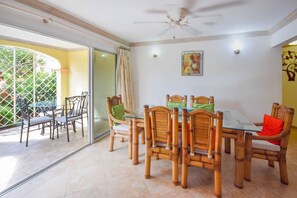 Un comedor con mesa de vidrio, sillas de madera, ventilador de techo y vista a un jardín a través de puertas de cristal.