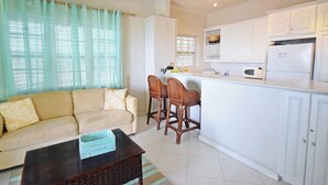 Una sala de estar con sofá y mesa de centro, una cocina con armarios blancos y un refrigerador, y un ventanal con cortinas traslúcidas.