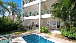 Un área de piscina con un edificio de dos pisos, mobiliario exterior y plantas tropicales.