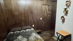 Un dormitorio con techo de madera, cama con ropa de cama estampada, un taburete de madera pequeño y una decoración en la pared.