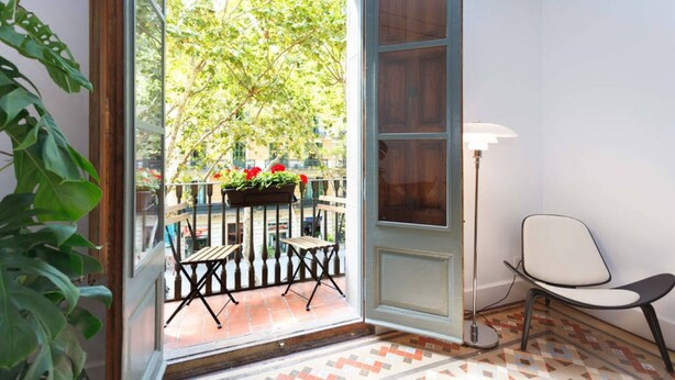 Una habitación con un ventanal grande, un pequeño balcón con mesa y sillas, una planta en maceta y una lámpara de pie.