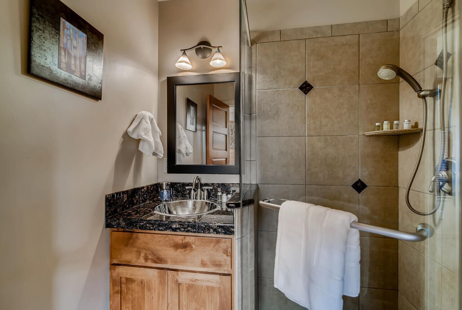 A bathroom with a granite countertop sink, a wooden vanity, a large mirror, and a shower with a handheld showerhead.