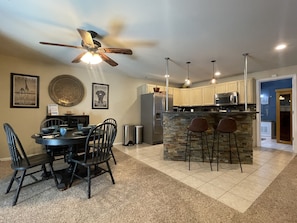 A modern kitchen with a central island, a dining area with chairs, a ceiling fan, and a stone backsplash.