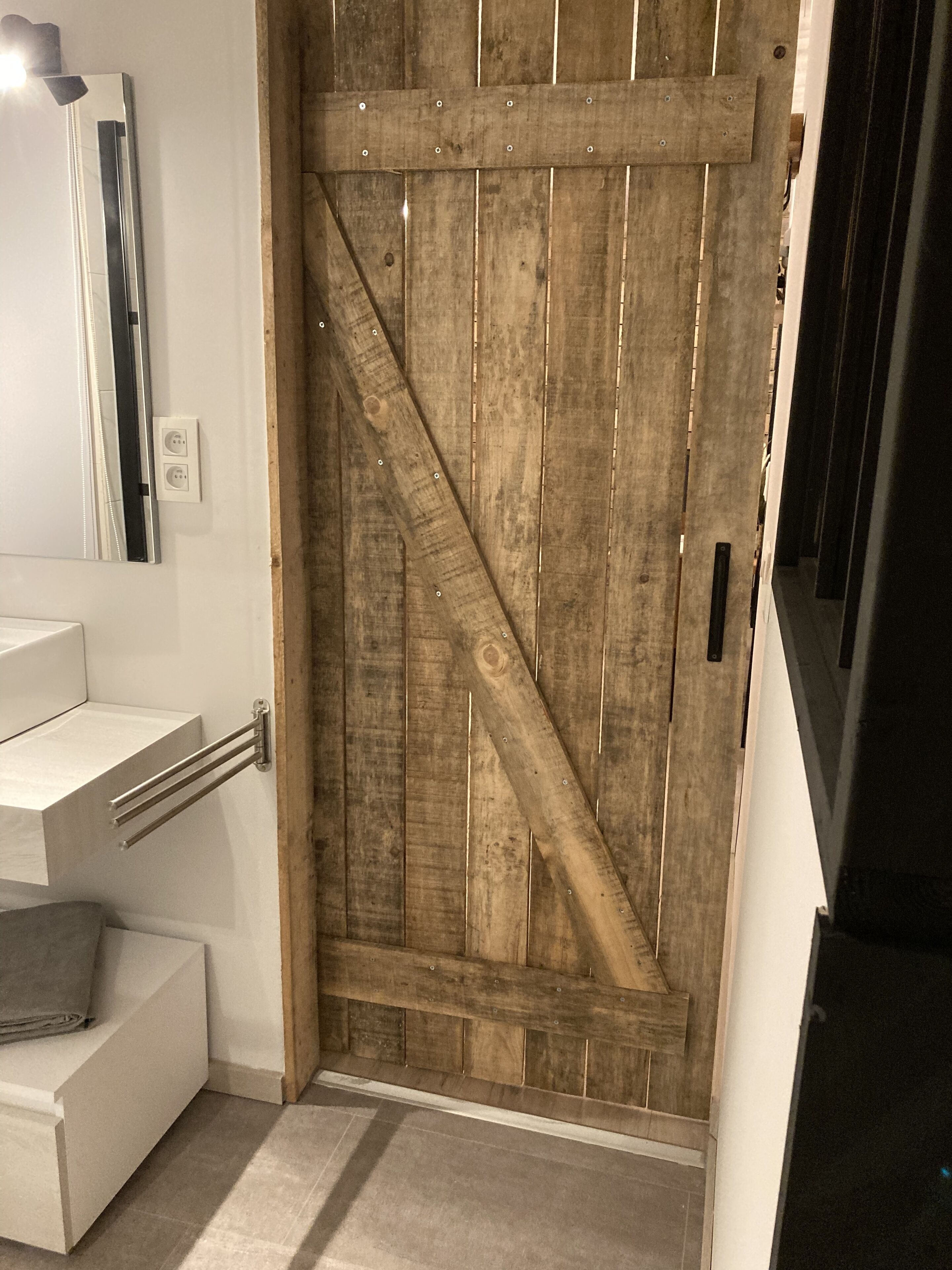 Une porte en bois avec une traverse métallique diagonale, un mur blanc orné d’un miroir et un plan de travail blanc muni d’un porte-serviettes.
