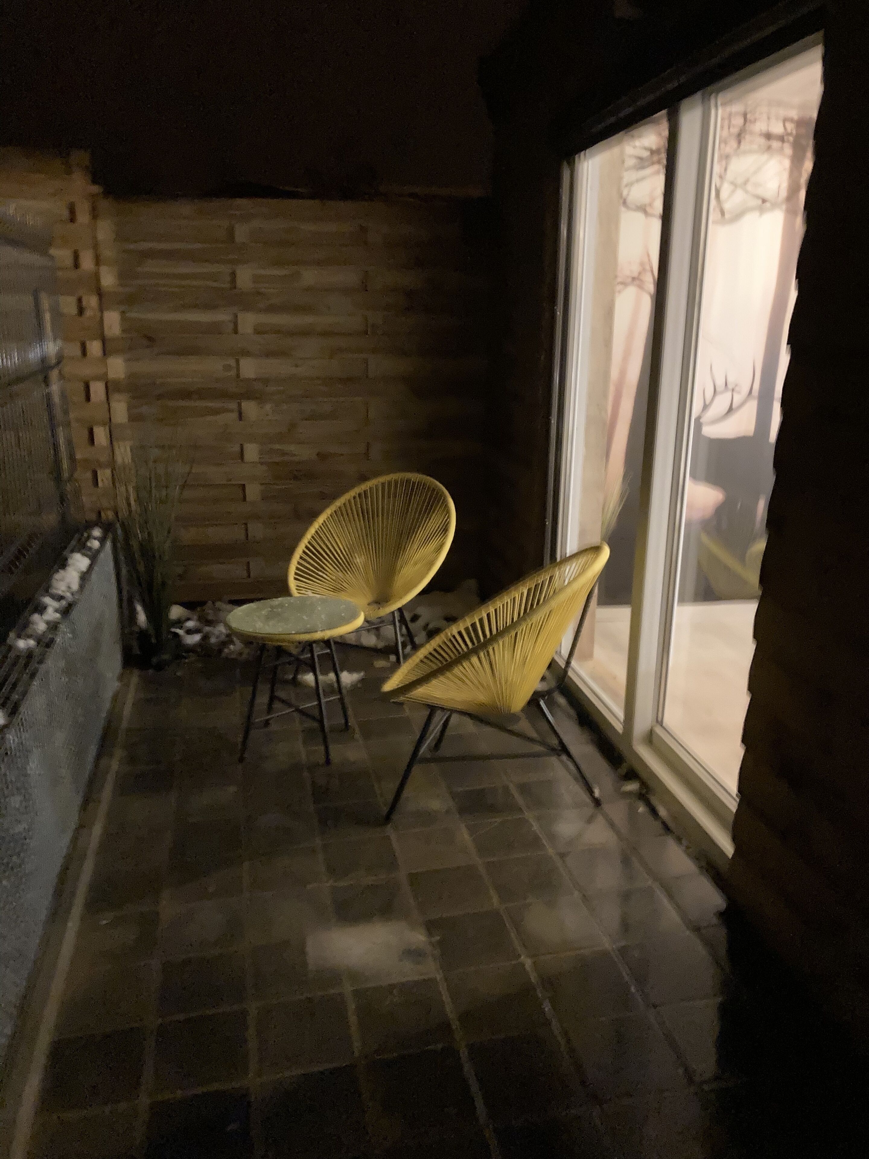 A patio with two yellow chairs and a small table, a brick wall, and a glass door leading outside.
