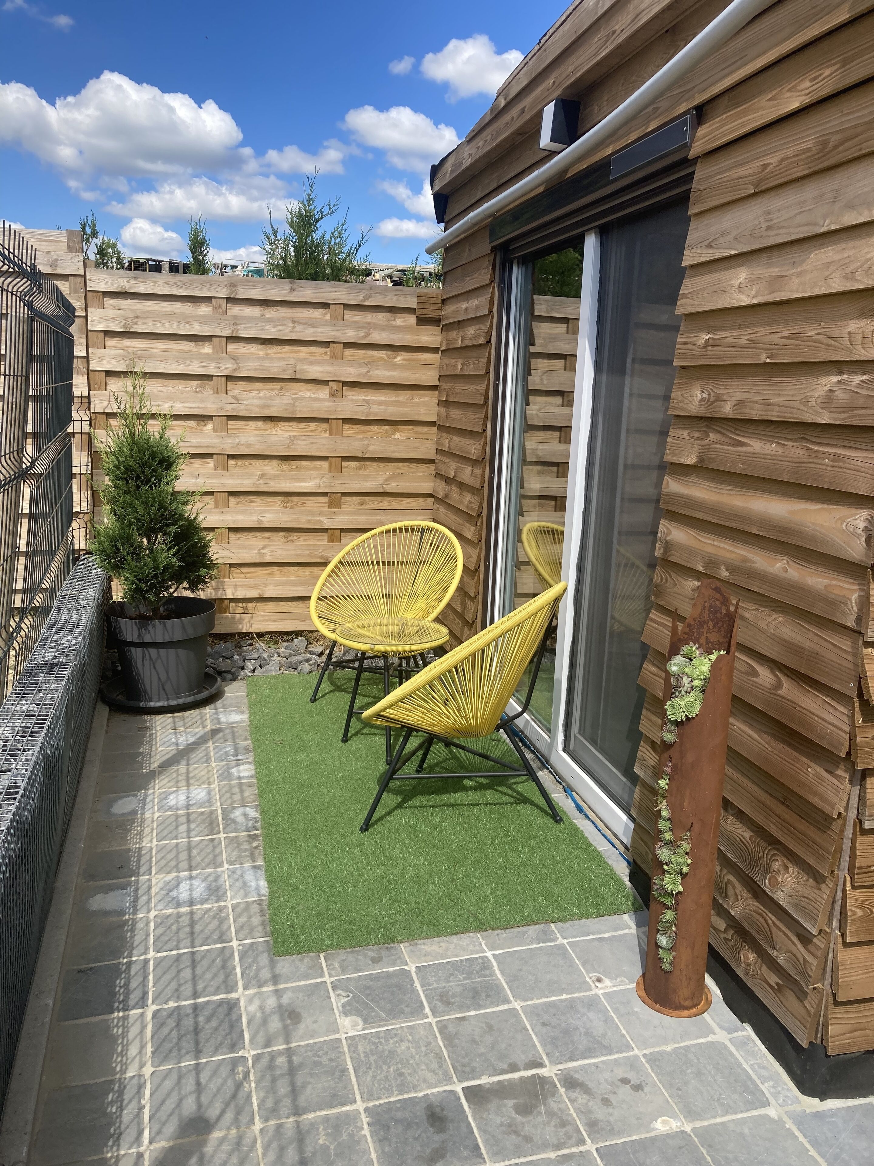 A patio with two yellow chairs, a small potted plant, and a wooden fence.