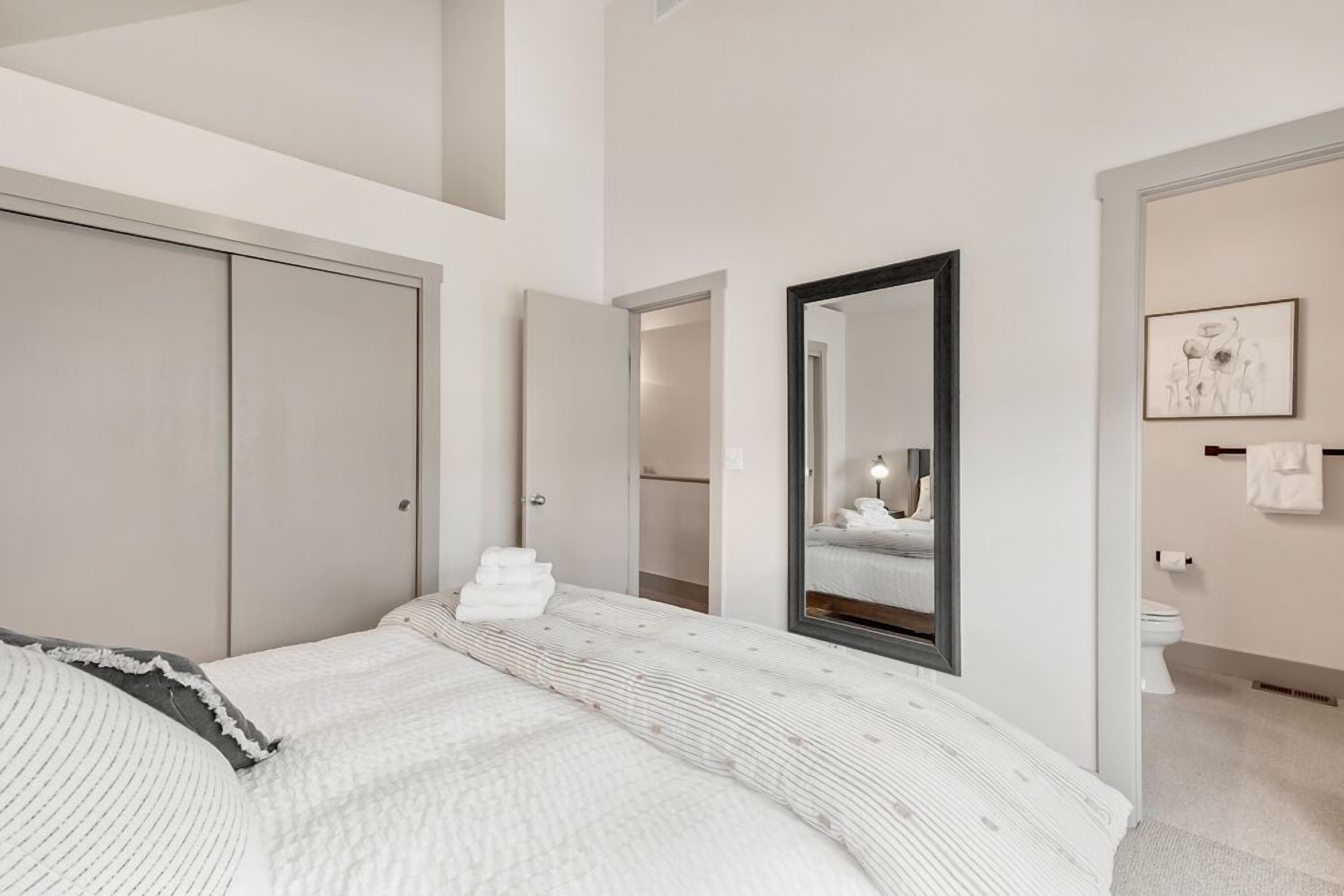 Una camera da letto moderna con un letto grande, un armadio, uno specchio e un bagno visibile attraverso una porta aperta.