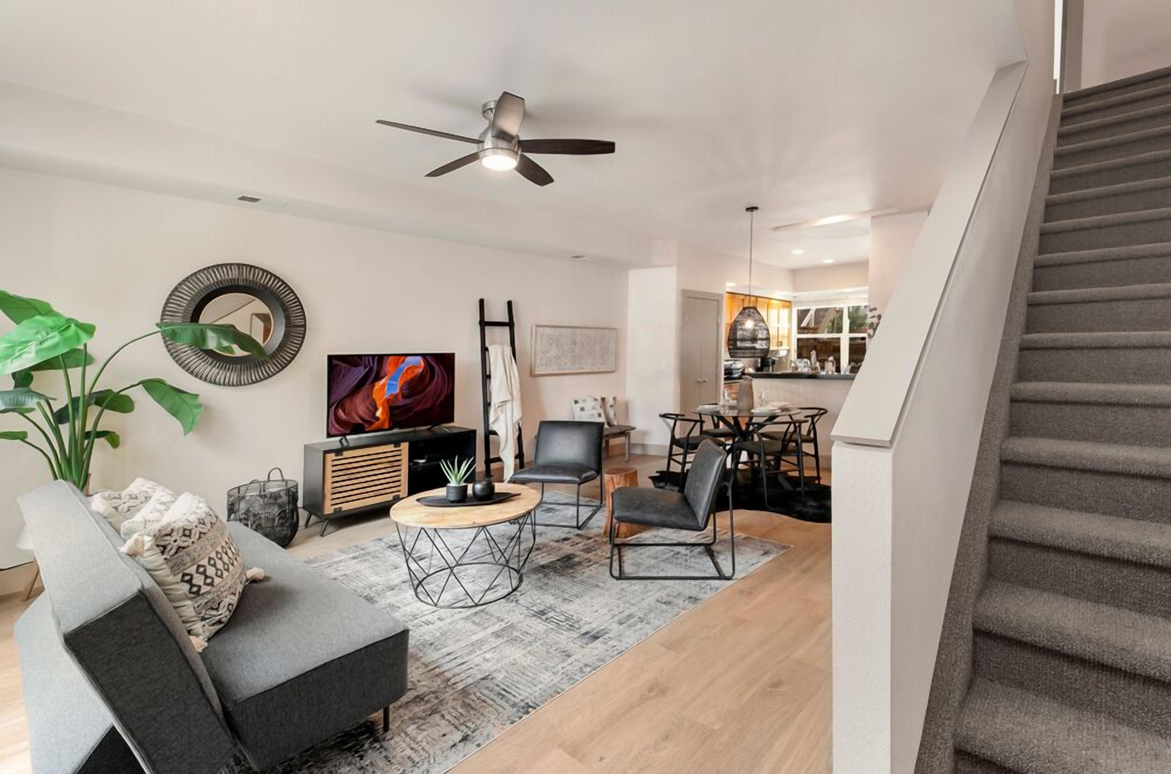 A modern living room with a sofa, coffee table, and TV. There's a staircase to the right and a kitchen in the background.