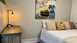 A bedroom with a wooden nightstand, a lamp, a bed with patterned pillows, and a colorful abstract painting on the wall.