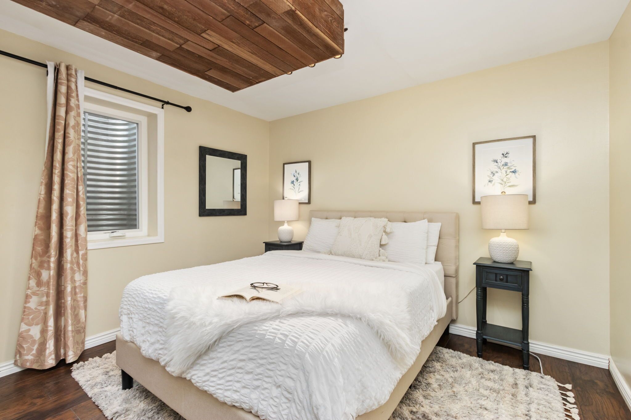 A bedroom with a large bed, two bedside tables with lamps, a window with blinds, and a wooden ceiling.