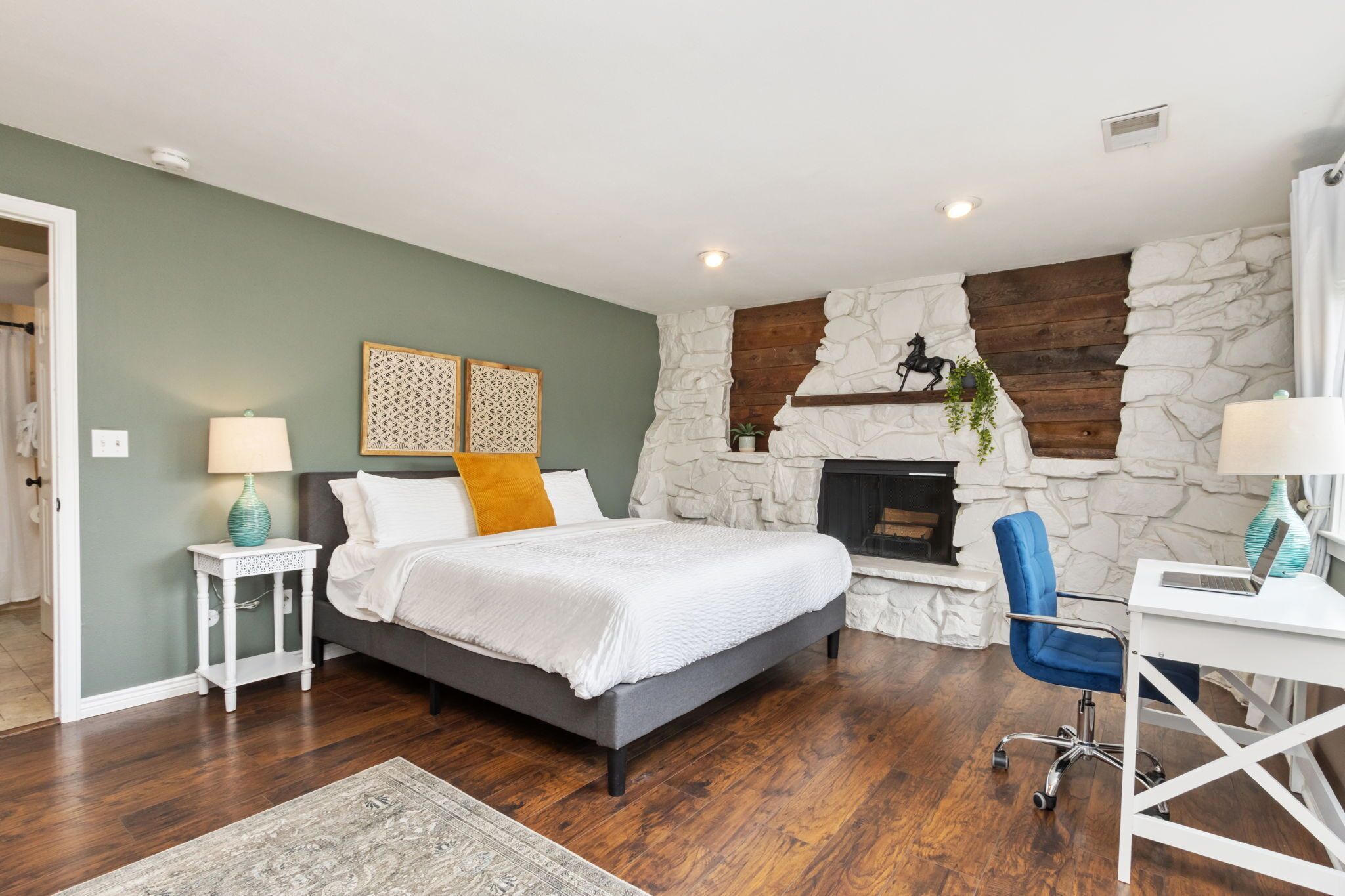 Un dormitorio con chimenea de piedra, una cama con ropa de cama blanca, un escritorio con una computadora portátil y una silla azul.