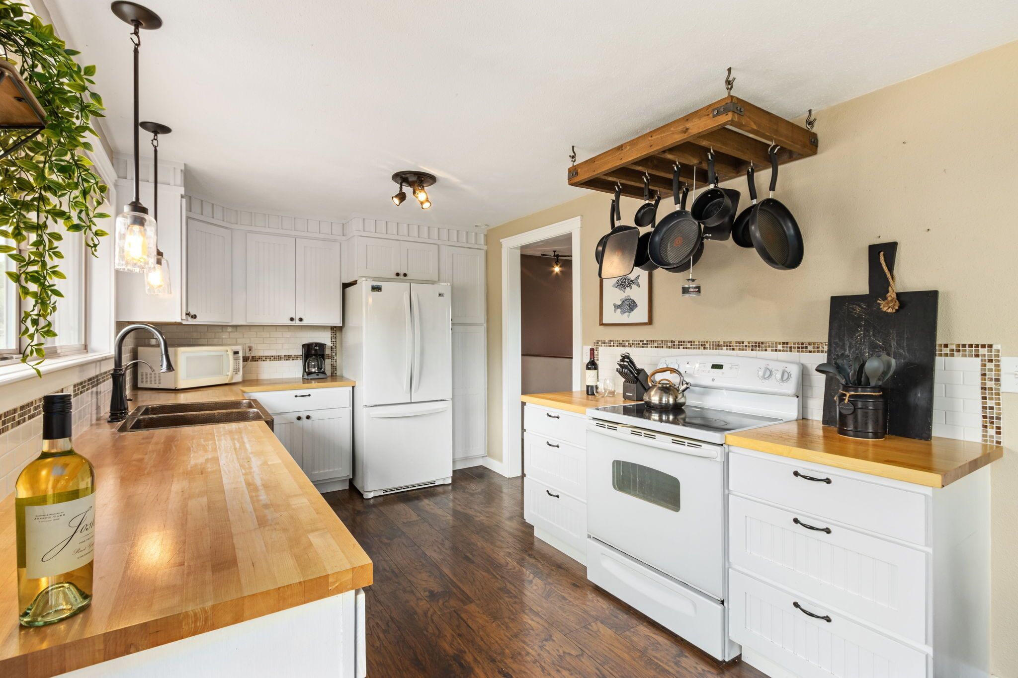 Une cuisine avec des meubles blancs, un plan de travail en bois, un réfrigérateur blanc, une cuisinière et des casseroles suspendues.