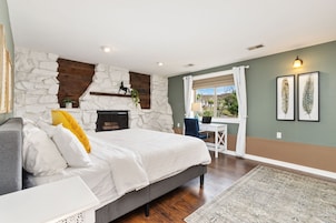 A bedroom with a stone wall, a bed with white bedding, a desk, and a view of the outdoors.