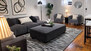 Sala moderna com sofá cinza, um banquinho preto, um tapete listrado e duas poltronas. Há quadros abstratos na parede, um abajur e um vaso com planta.