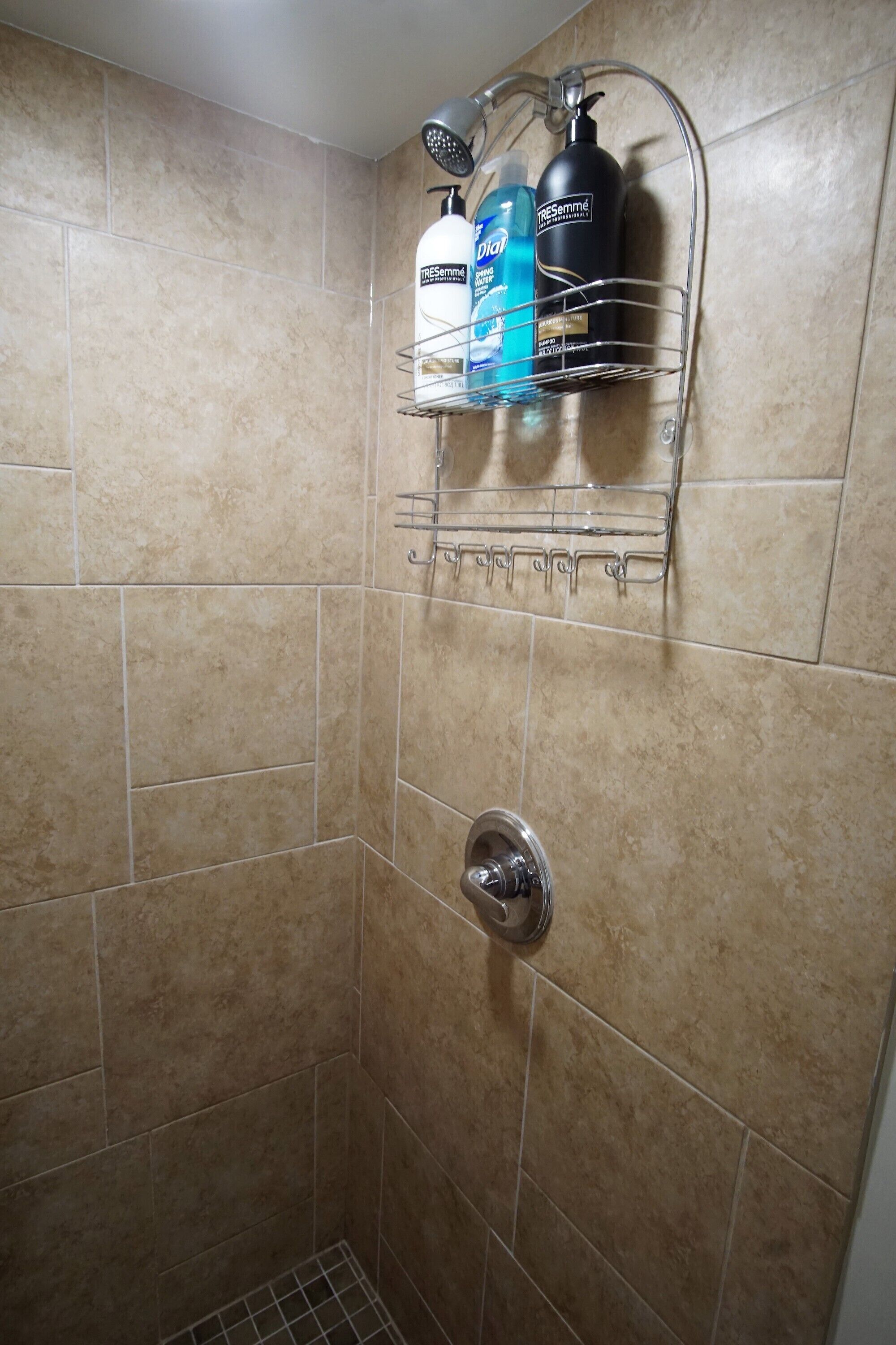 Zona de ducha con azulejos color crema, cabezal de ducha y un estante metálico con artículos de aseo.