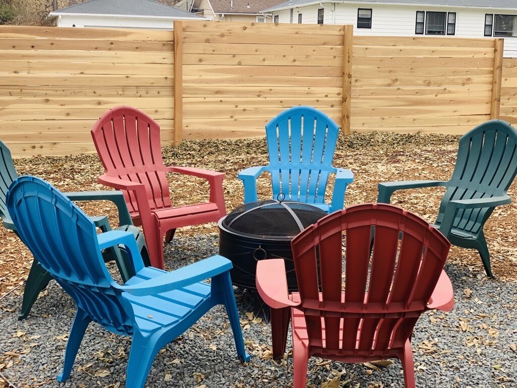 Un ensemble de fauteuils Adirondack de différentes couleurs est disposé autour d’un foyer dans un espace extérieur.